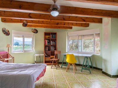 Bright bedroom featuring exposed wooden beams and light green walls. It has patterned floor tiles, windows with natural views, and a study area.