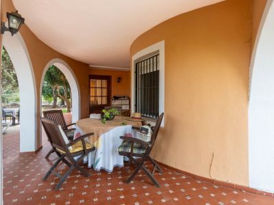 Covered terrace featuring reddish-brown tiled flooring, white arched openings to the garden, and an orange curved wall. Furnished with a round table and wooden chairs.