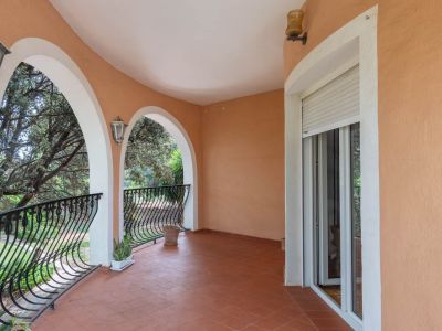 Covered terrace with terracotta tiles, featuring elegant arched openings with white trim and decorative wrought iron railings. Offers views of surrounding trees and includes a sliding glass door.