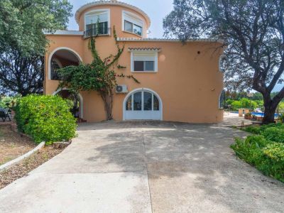 Exterior view of the peach-colored villa, featuring its rounded architectural section, balcony, and main entrance. A large concrete driveway and mature garden with a pool are visible.