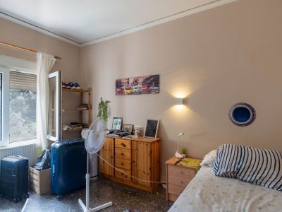 Functional bedroom featuring a single bed, wooden nightstand, and chest of drawers, illuminated by natural light from the window and a wall light. Dark, speckled flooring.