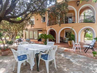 House exterior featuring a peach-toned facade with ground-floor arches and upper balconies. A large, leafy tree shades the terrace, furnished with an outdoor table and a hammock.