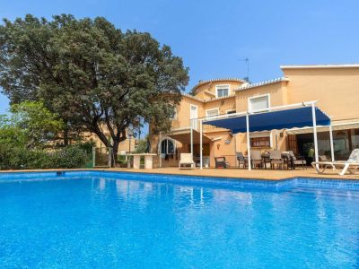 Large blue-tiled swimming pool with an expansive adjacent terrace featuring an outdoor dining area under a blue pergola. The ochre-colored house facade is framed by a mature tree.