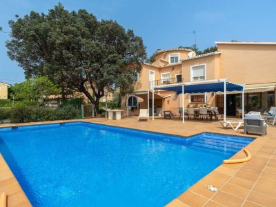 Large rectangular swimming pool with terracotta tiled surround and spacious deck. Features an outdoor dining area under a blue pergola and the peach-toned facade of the house.