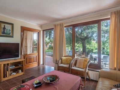 Living room featuring tiled flooring and large wooden-framed windows overlooking the garden. Includes a leather sofa, wicker armchairs, and direct outdoor access.