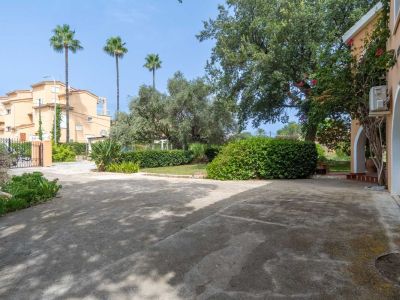 Spacious concrete private parking area, bordered by a Mediterranean garden featuring palm trees, olive trees, and lush shrubs. Part of the chalet's arched facade is visible.