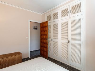 This bedroom features a spacious built-in wardrobe with white louvered doors, a dark wooden entrance door, and a bed. A wicker chest adds a touch of warmth.
