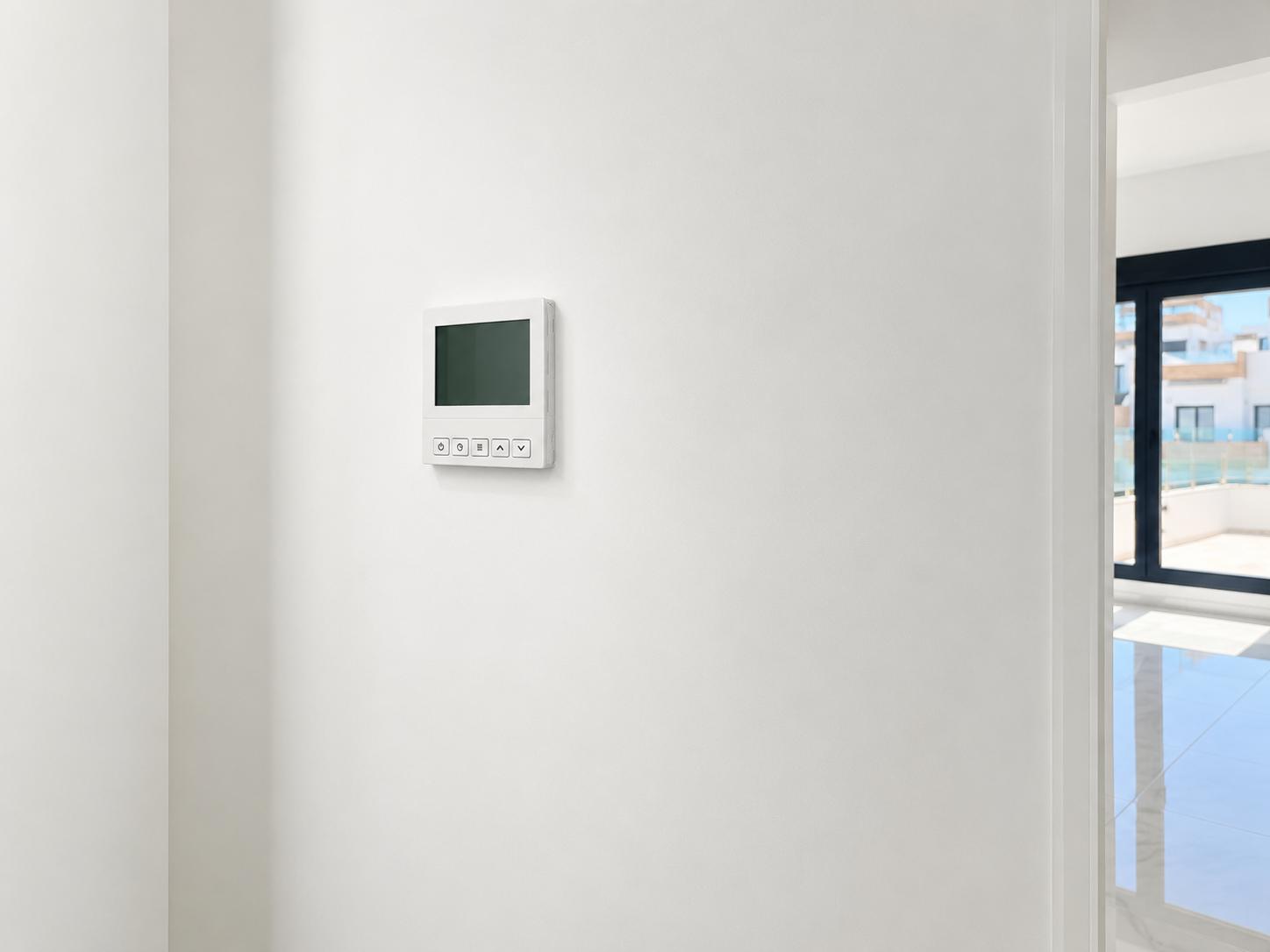 A sleek digital climate control panel is mounted on a pristine white wall, offering a glimpse into a bright adjacent room with tiled flooring and an outdoor terrace view.