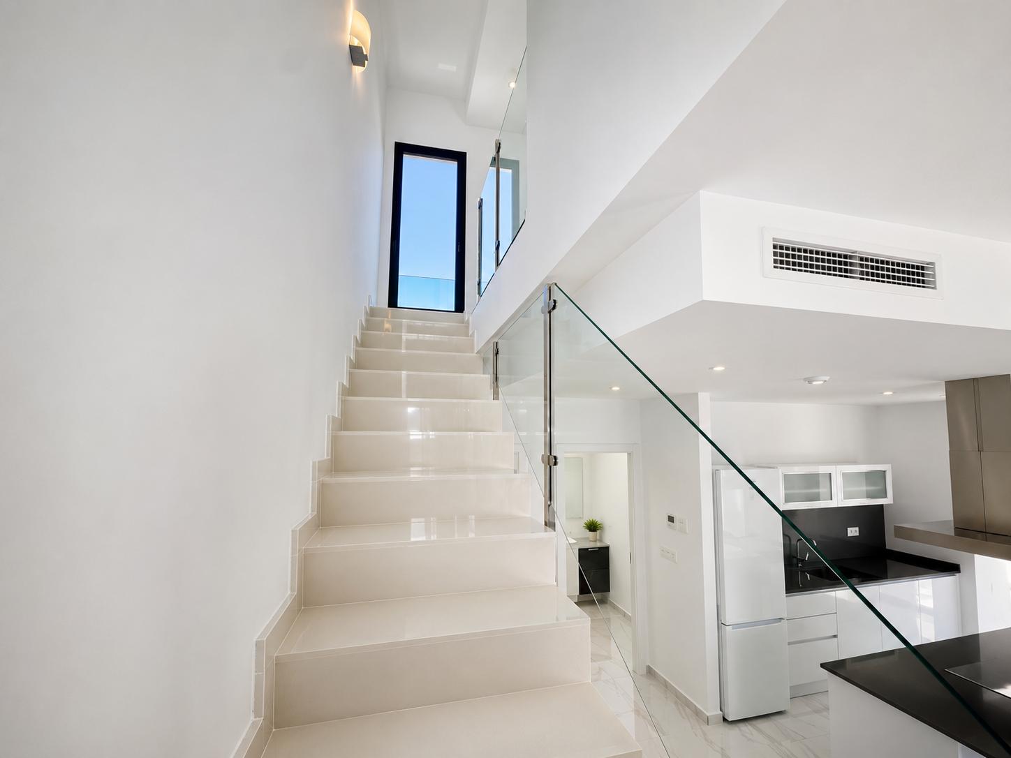Bright staircase with light-toned tiles and a glass balustrade, leading to an exterior door. Below, a modern kitchen with dark countertops and white appliances is visible.