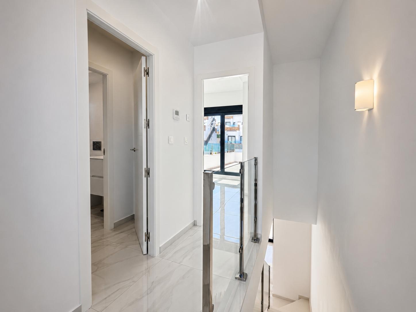 Bright upper-floor hallway featuring polished marble-look floor tiles and a modern glass and steel staircase railing. A doorway leads to a bathroom, another to an exterior balcony with natural light.