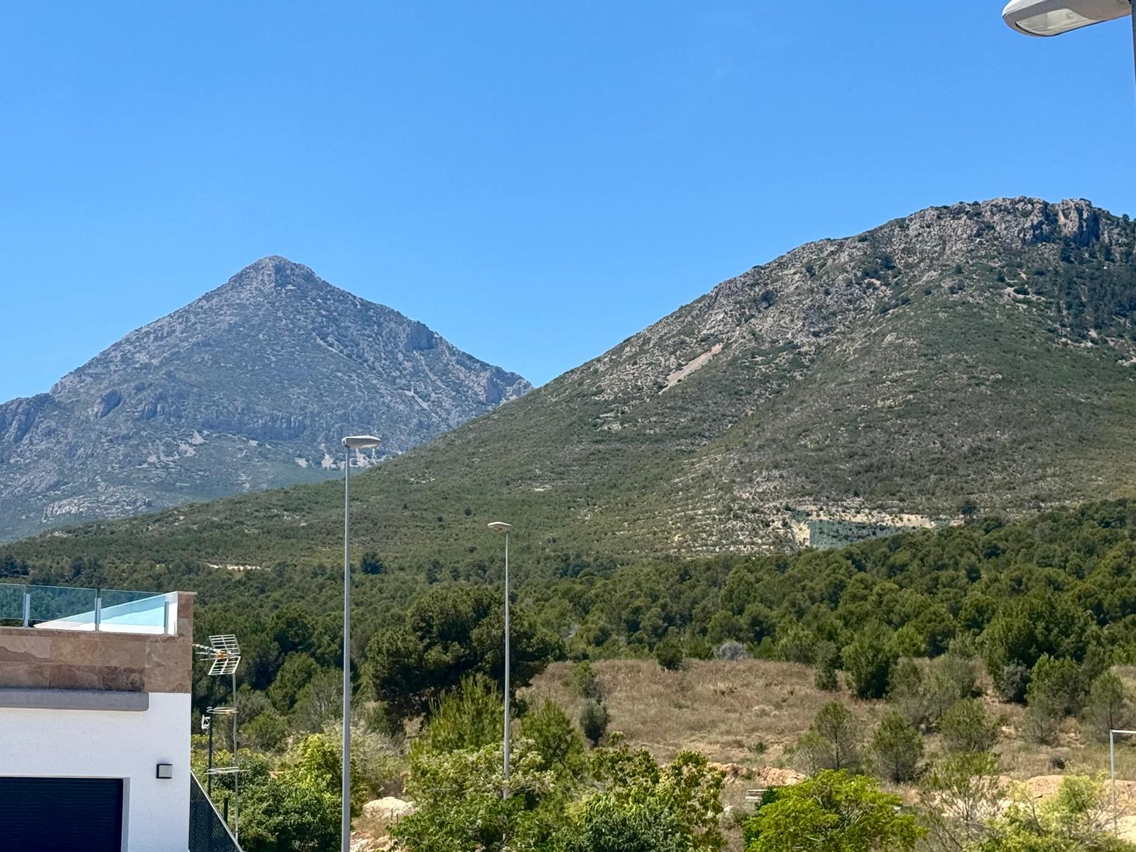 Clear views of two distinct rocky mountains and lush Mediterranean vegetation, complemented by a modern terrace with a glass railing in the foreground.