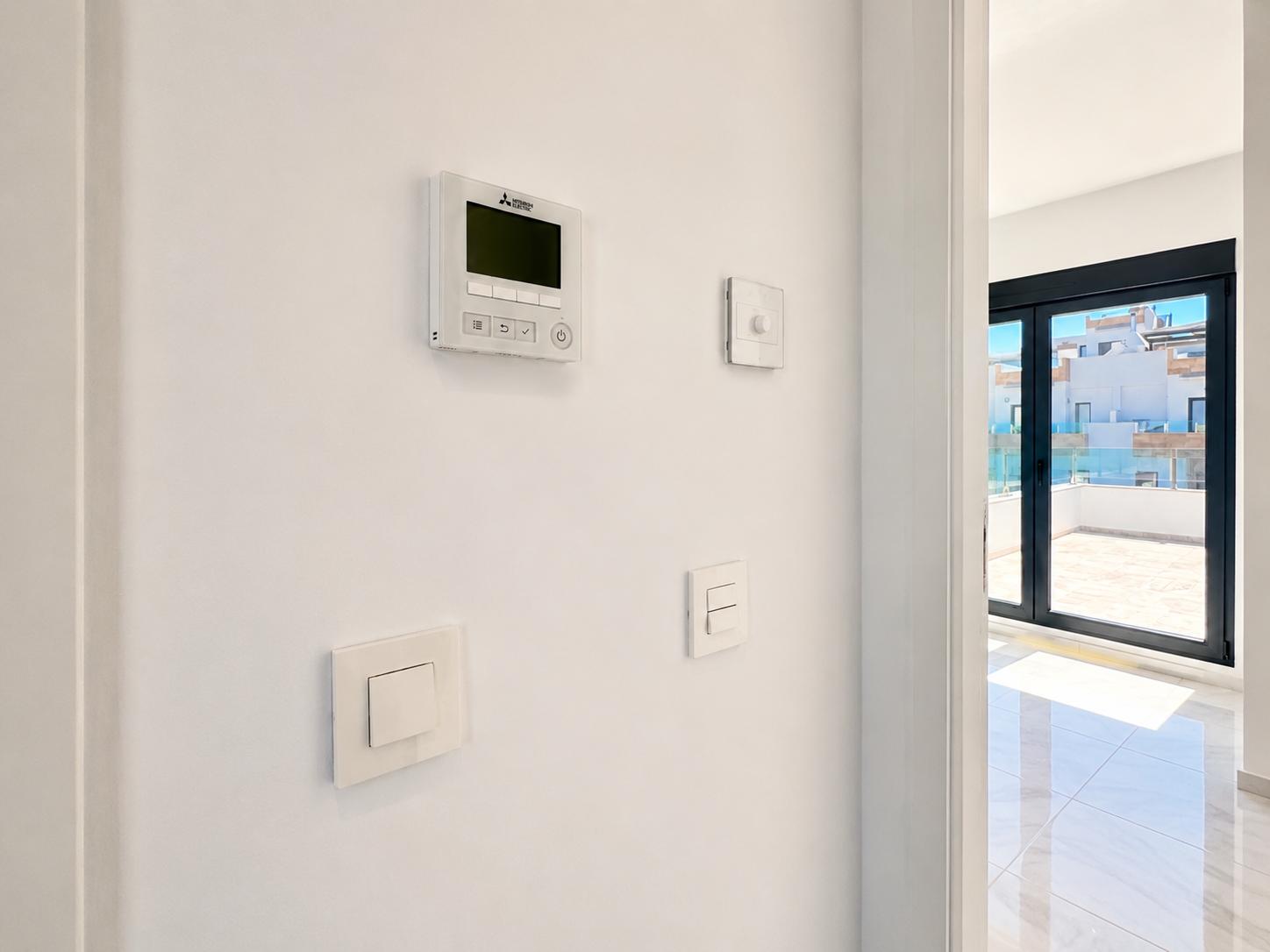 Mitsubishi air conditioning control panel and modern light switches on a clean white wall, adjacent to large sliding doors opening onto a tiled terrace with glass railings.