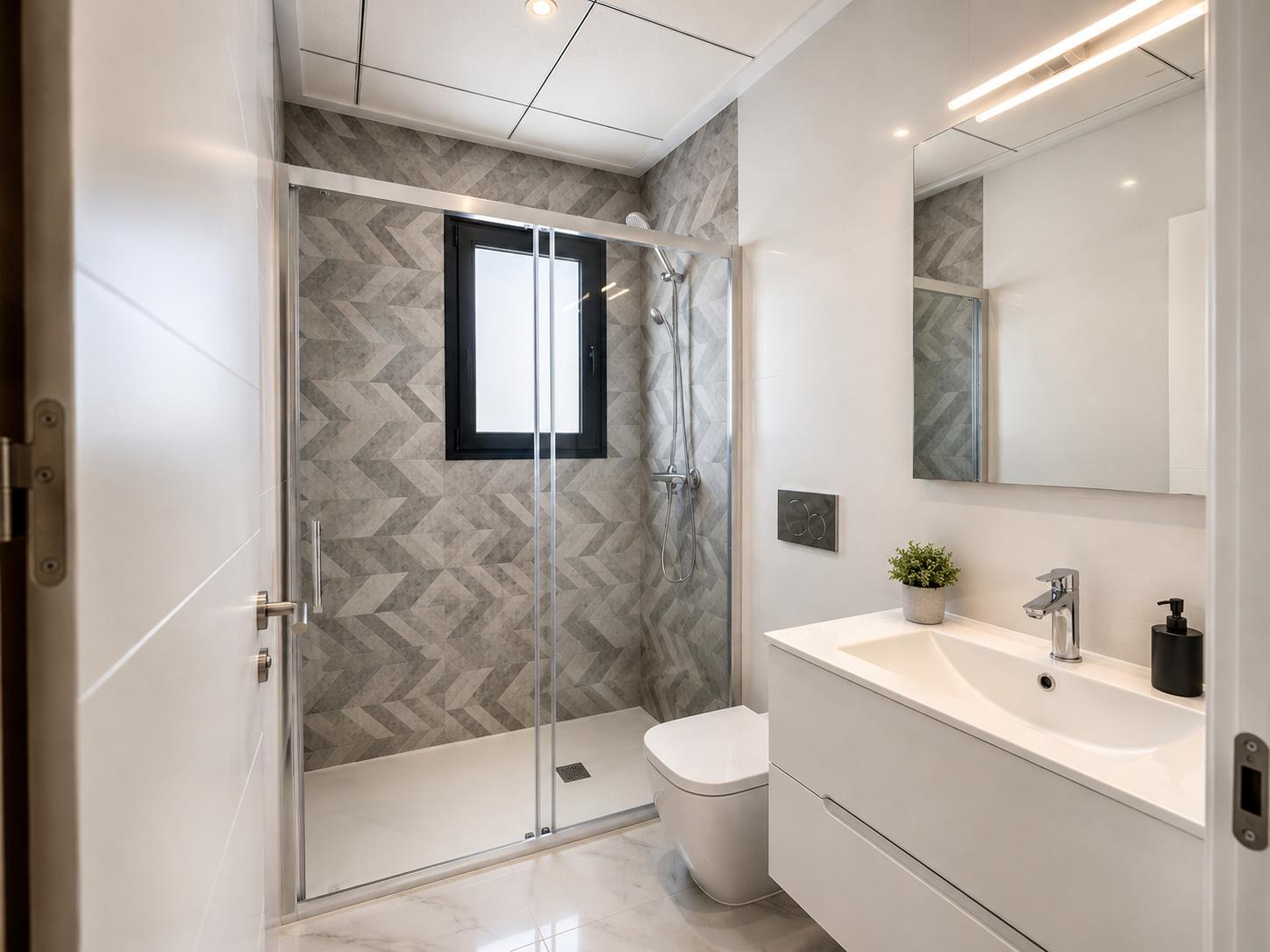 Modern bathroom featuring a glass-screen shower with geometric grey tiles and a window. White vanity, LED-lit mirror, and marble-look floor tiles.