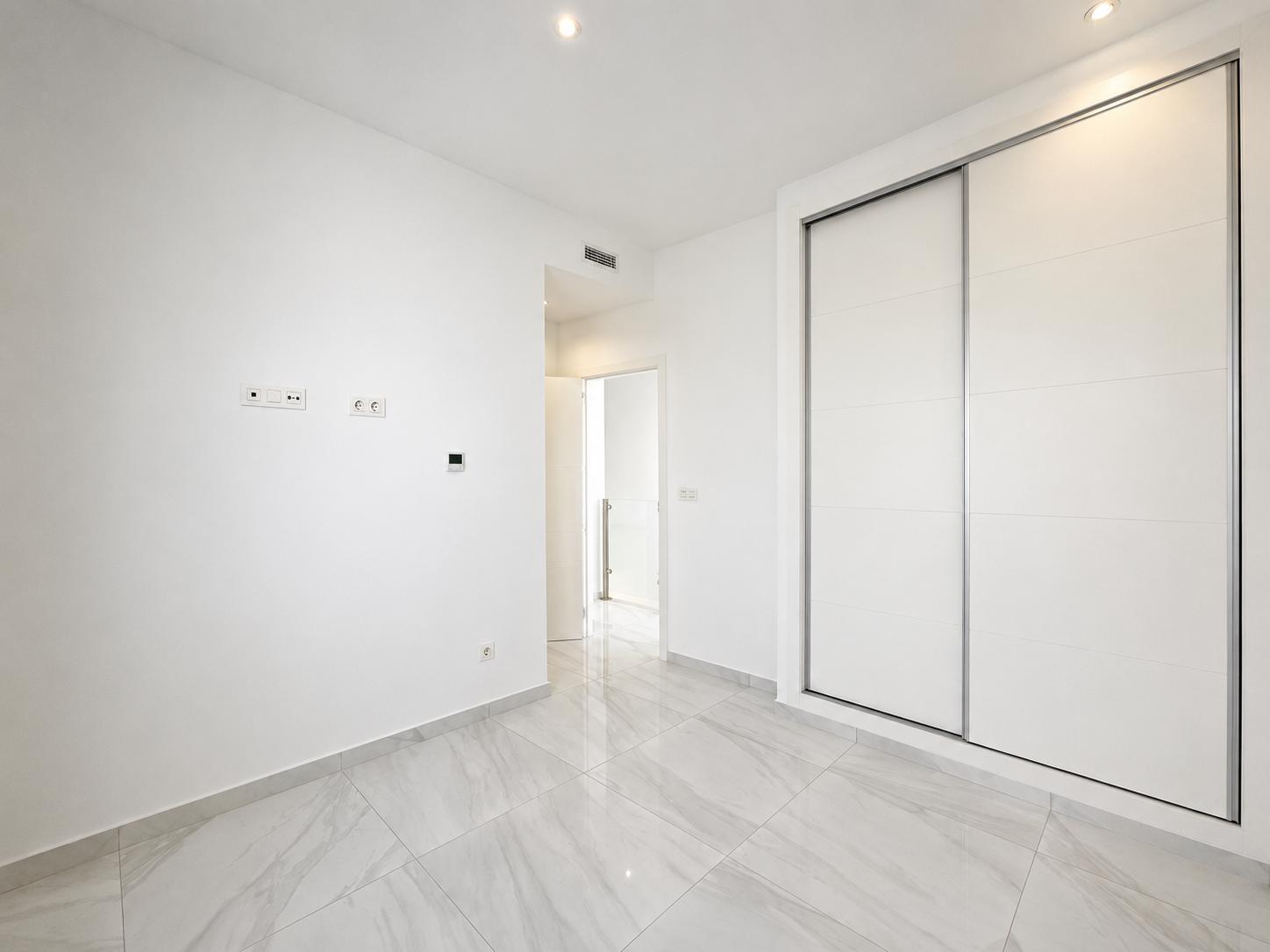 Modern room featuring large white built-in sliding wardrobes, light grey marble-effect floor tiles, and recessed ceiling lights. A clean, functional design.