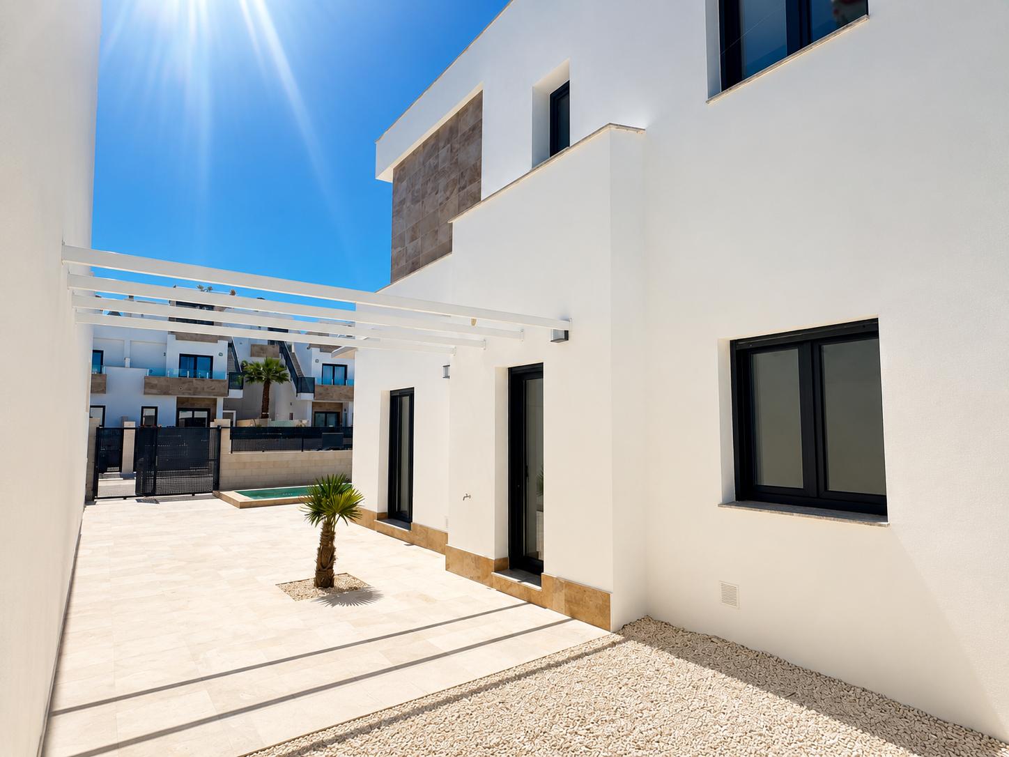 Modern villa exterior featuring a white facade with stone accents. Light-tiled terrace, a small palm tree, white pergola, and dark-framed windows under bright sun.