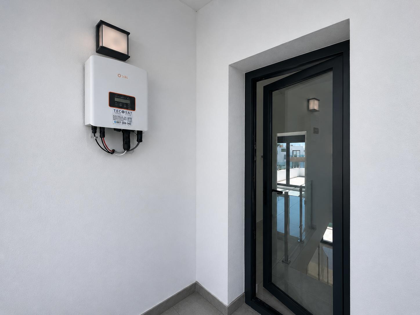 Solis solar inverter mounted on a white wall with an outdoor light fixture. A partially open black-framed glass door reveals an interior staircase with a railing.