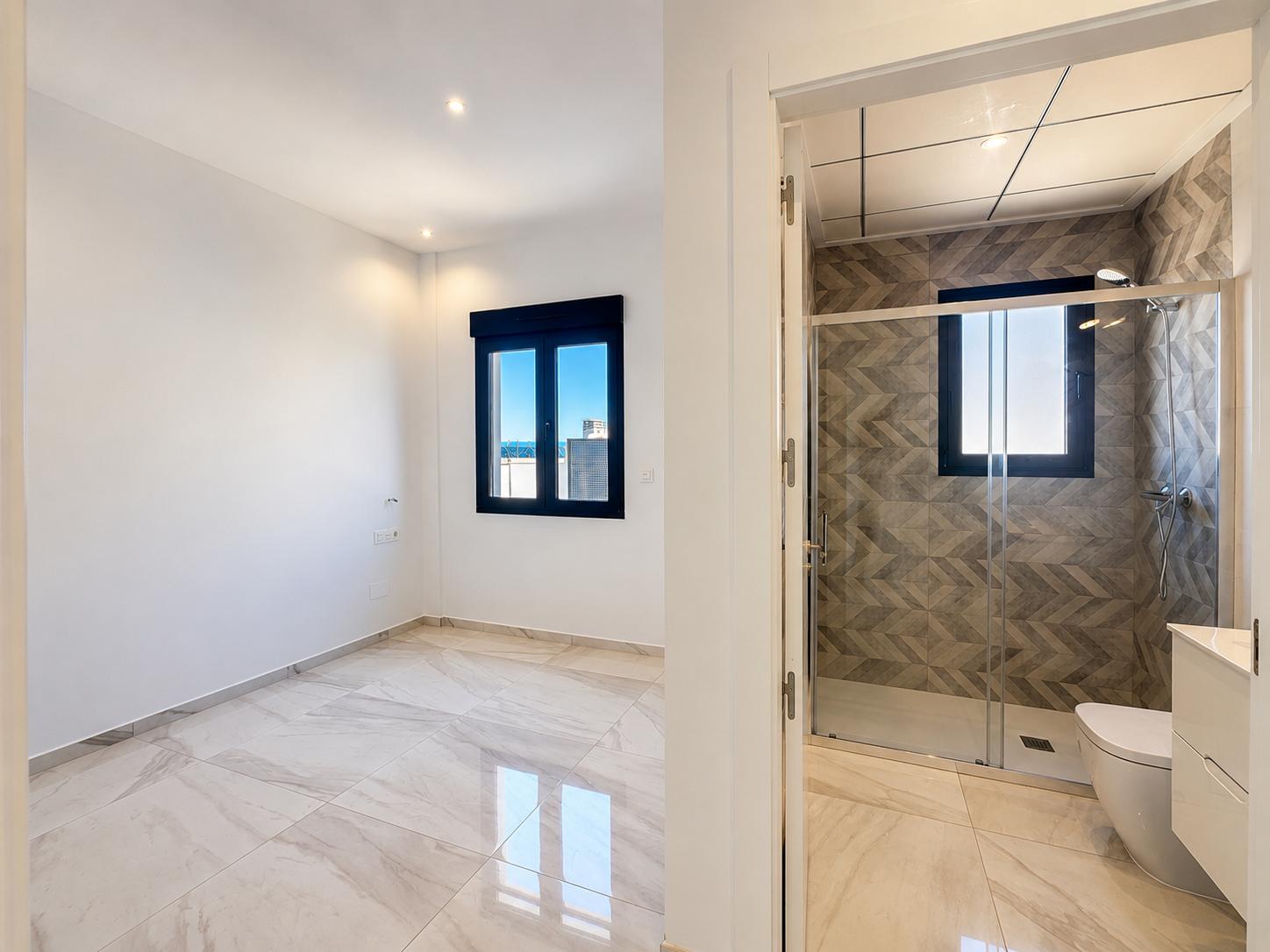 Spacious bedroom with light marble-effect flooring and a dark-framed window offering distant views, adjacent to an en-suite bathroom featuring geometric tiled shower walls.
