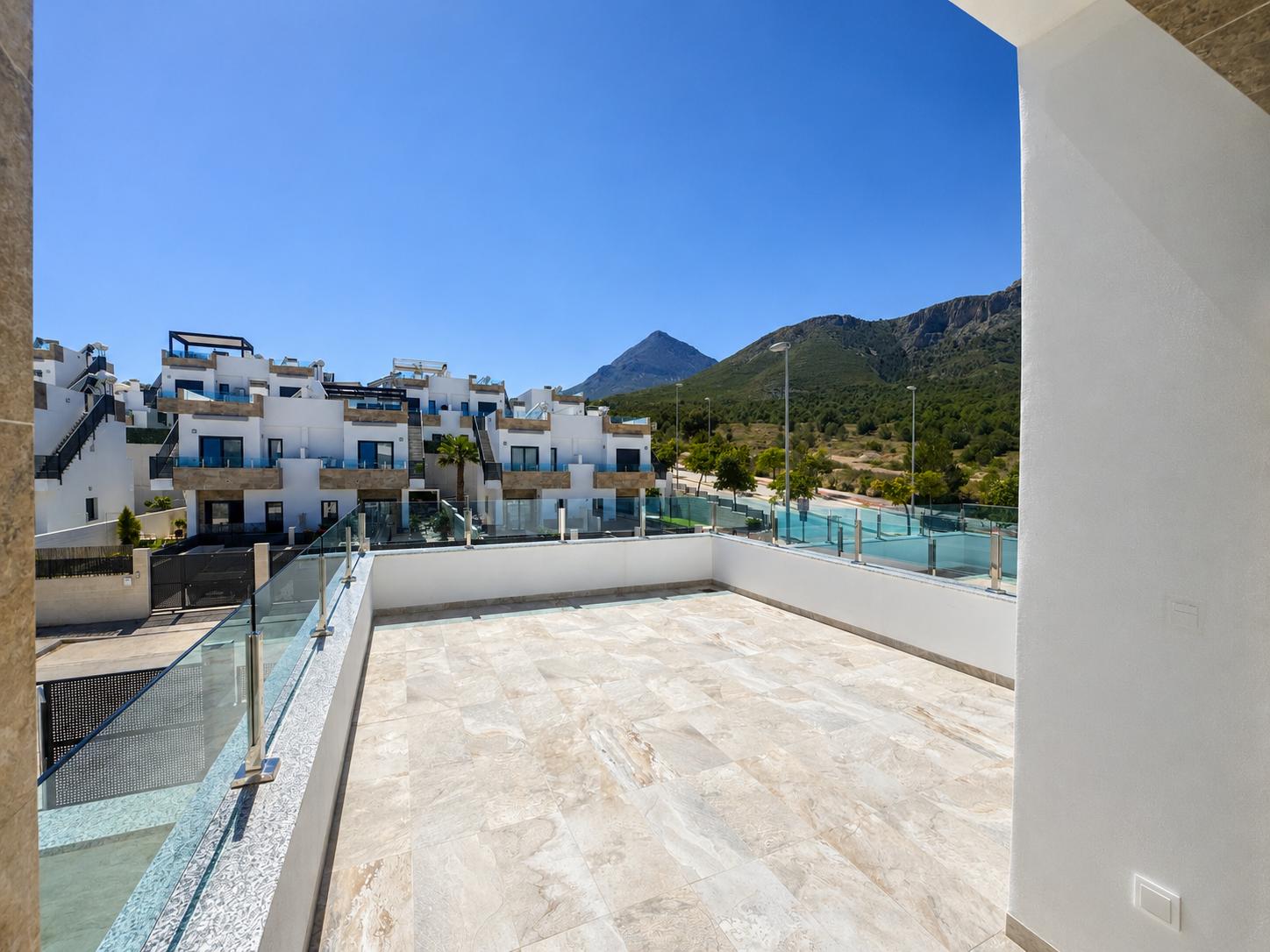 Spacious outdoor terrace featuring light-toned tiled flooring, glass railing, and views of modern residential buildings and tree-covered mountains under a clear blue sky.