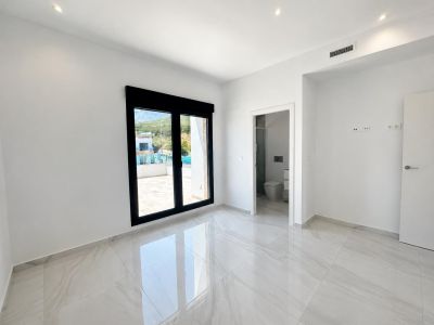 Bright bedroom featuring glossy white tiled floors, a large black-framed sliding door to an outdoor terrace, and an en-suite bathroom entrance.