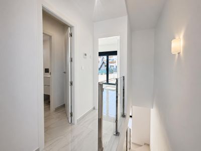 Bright upper-floor hallway featuring polished marble-look floor tiles and a modern glass and steel staircase railing. A doorway leads to a bathroom, another to an exterior balcony with natural light.