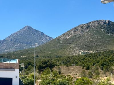Clear views of two distinct rocky mountains and lush Mediterranean vegetation, complemented by a modern terrace with a glass railing in the foreground.