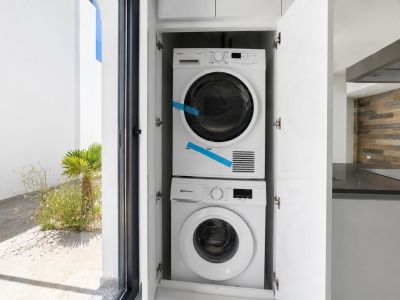 Integrated washer and dryer in white cabinetry, with direct access to a landscaped outdoor patio. A modern kitchen with wooden wall cladding and dark countertop is visible in the background.