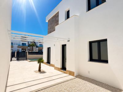 Modern villa exterior featuring a white facade with stone accents. Light-tiled terrace, a small palm tree, white pergola, and dark-framed windows under bright sun.