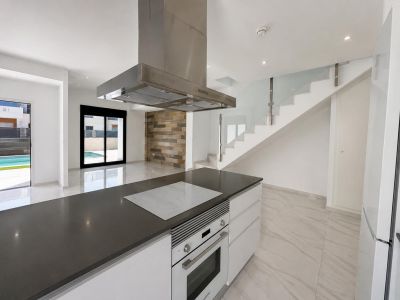 Open-plan kitchen with dark countertop island, integrated hob, and oven. The space connects to the living area and pool, featuring a glass staircase and stone accent wall.