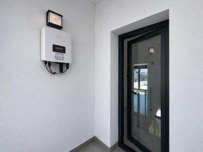 Solis solar inverter mounted on a white wall with an outdoor light fixture. A partially open black-framed glass door reveals an interior staircase with a railing.