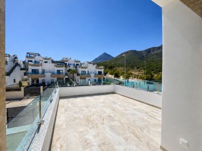 Spacious outdoor terrace featuring light-toned tiled flooring, glass railing, and views of modern residential buildings and tree-covered mountains under a clear blue sky.