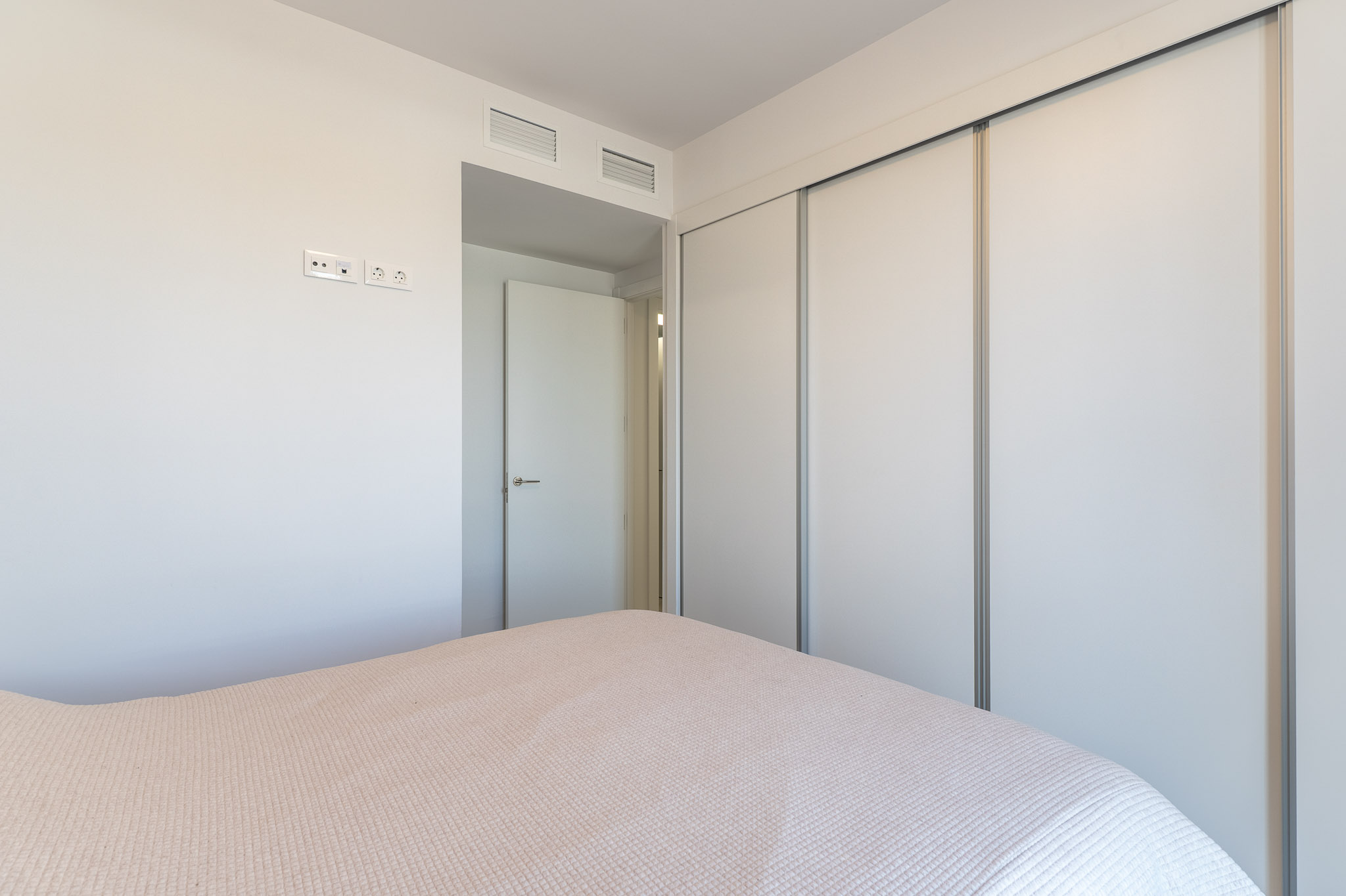 Bedroom view featuring a spacious built-in wardrobe with white sliding doors. The wall includes modern electrical outlets and integrated ventilation grilles above the adjacent doorway.