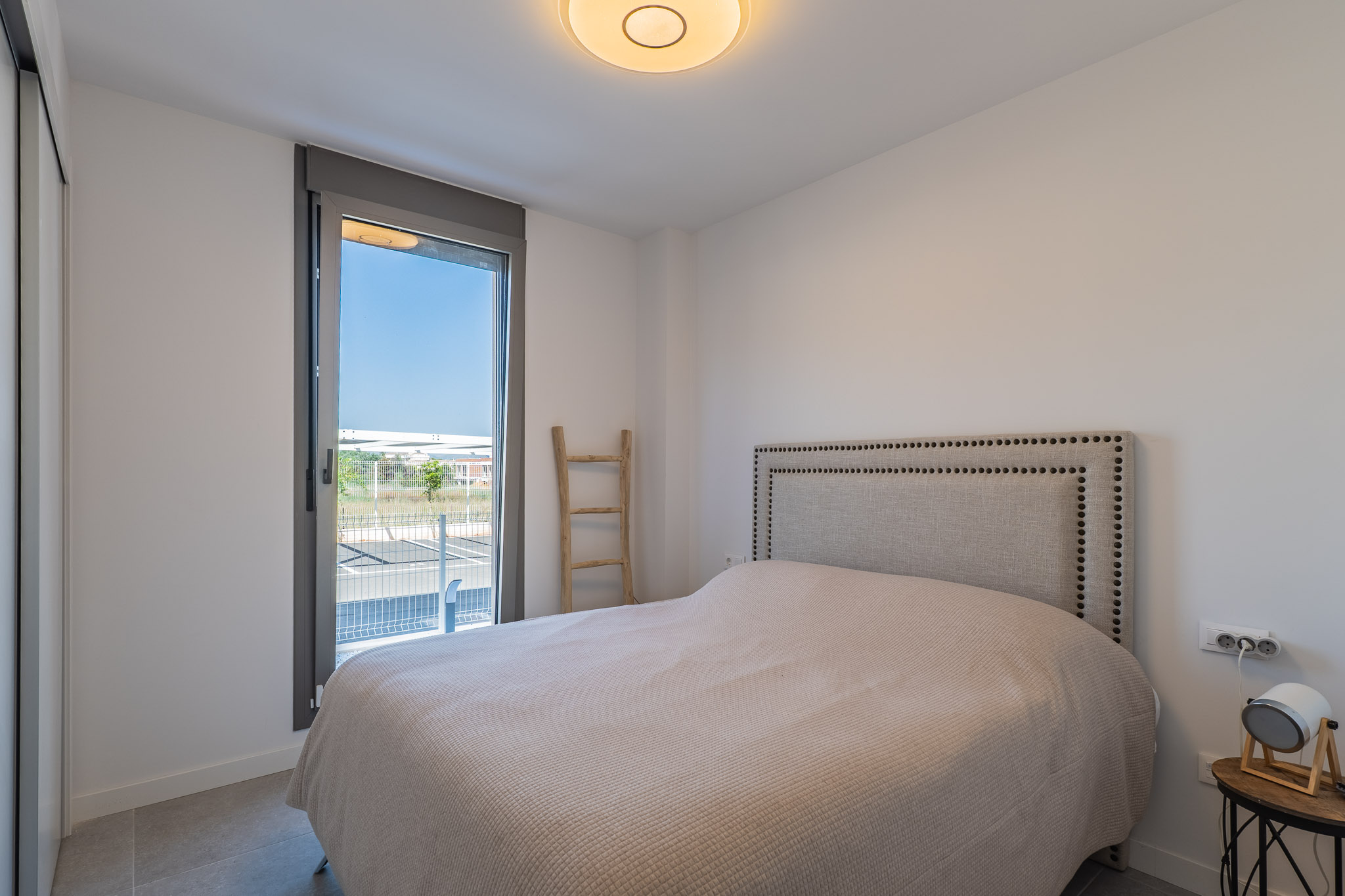 Bright bedroom featuring a bed with a studded fabric headboard and a built-in wardrobe. Large window offers outdoor access and natural light; grey tiled floor.