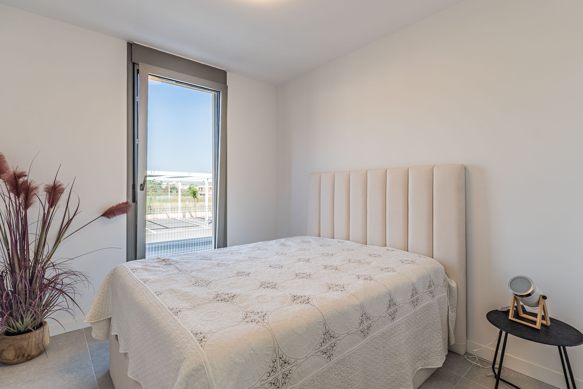 Bright bedroom featuring a bed with a tall, padded beige headboard. A large floor-to-ceiling window offers exterior views. Grey tiled floor and a decorative plant complete the space.