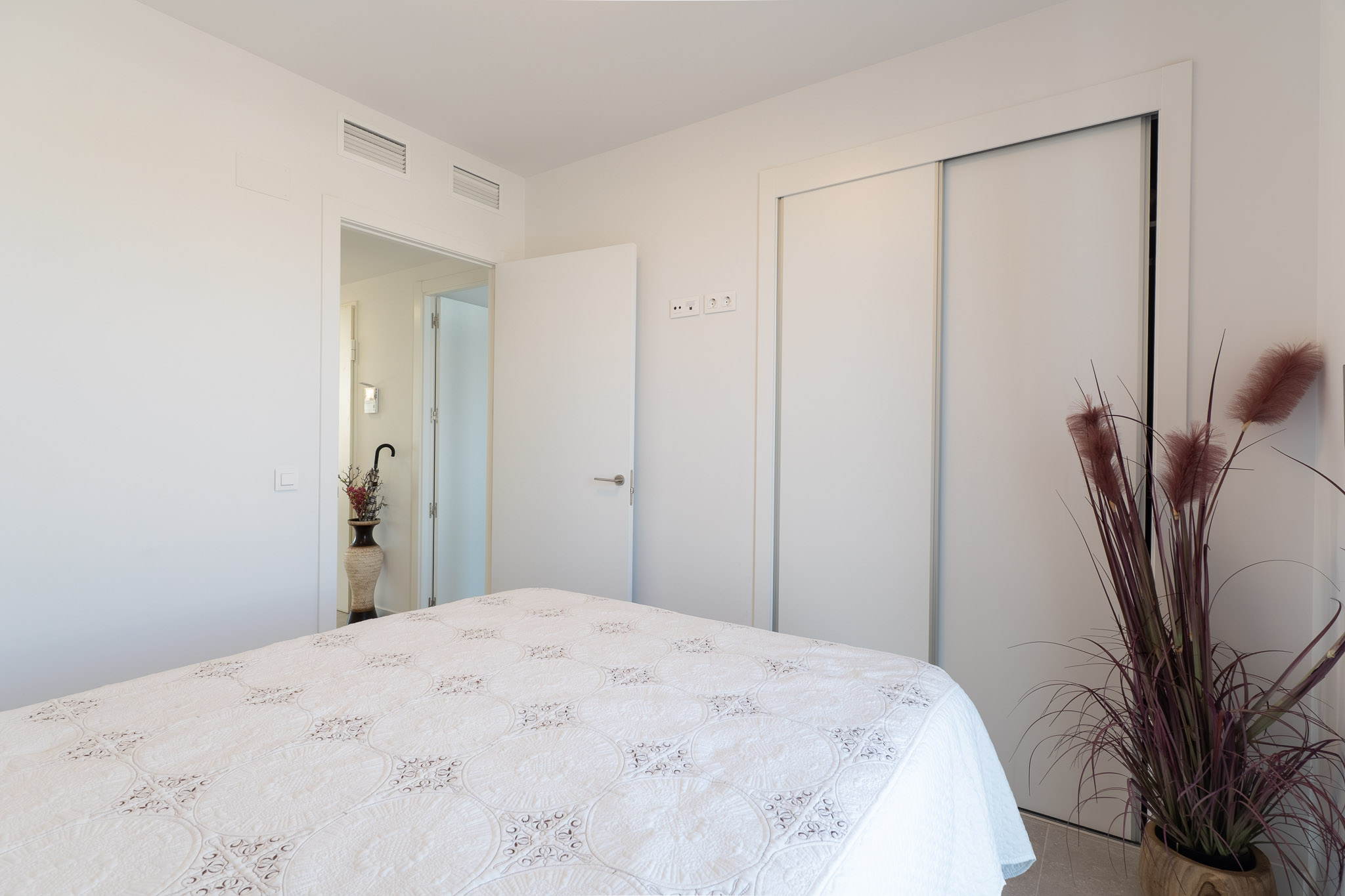 Bright bedroom featuring a bed with a white patterned quilt. Equipped with a built-in sliding wardrobe and integrated ceiling air conditioning vents. A decorative plant adds a touch of color.