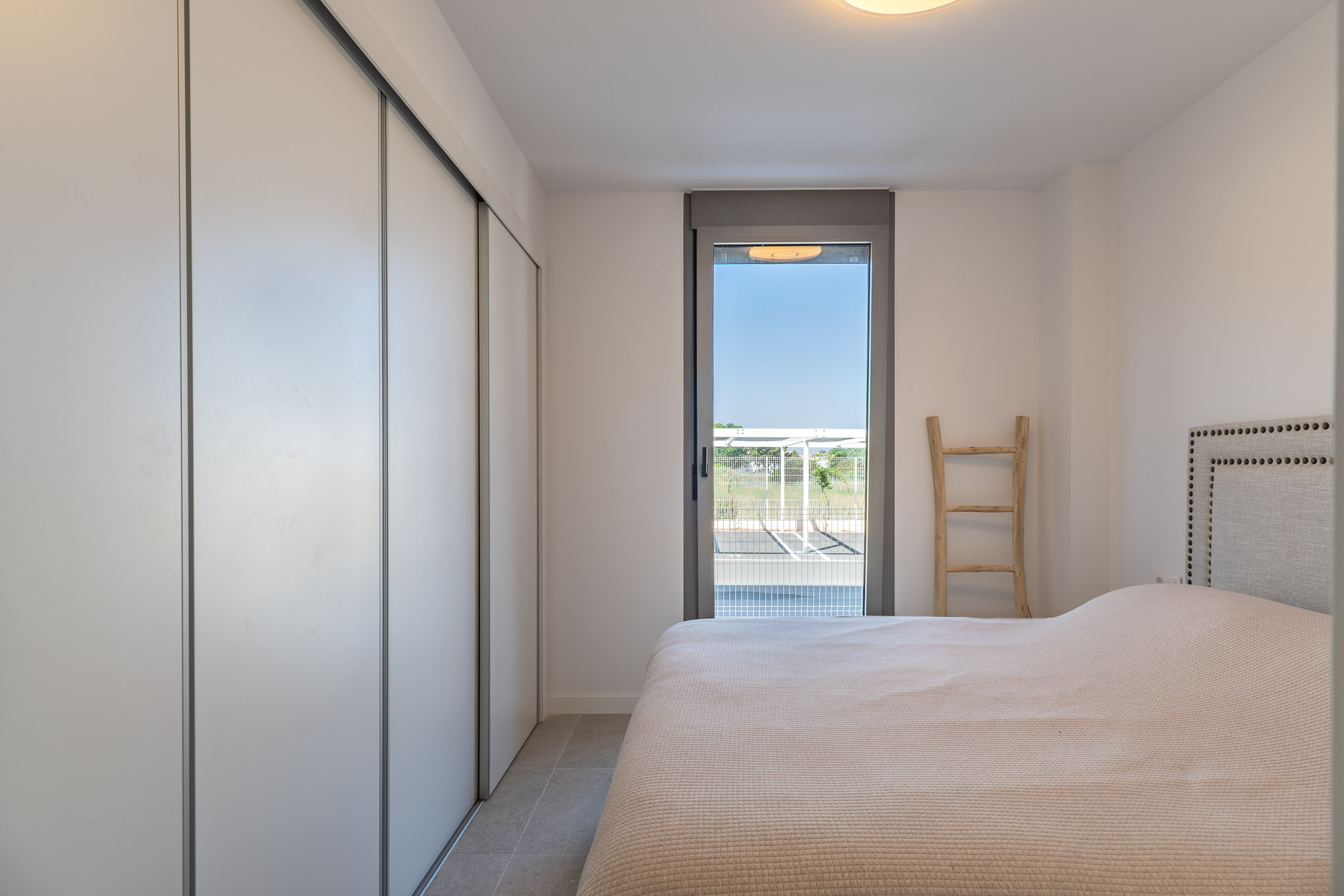 Bright bedroom featuring a built-in wardrobe with white sliding doors, tiled flooring, and direct access to the exterior through a glass door. Includes a bed with a studded headboard.