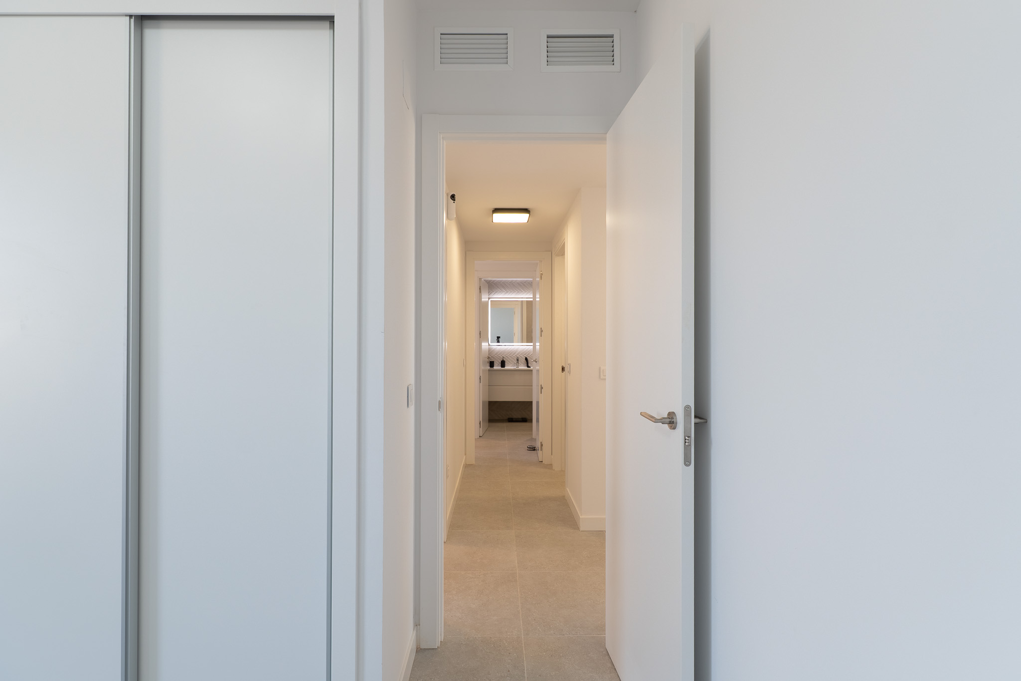 Bright hallway featuring a white sliding wardrobe and an open door. Light-toned floor tiles lead to a modern bathroom with a backlit mirror and contemporary vanity. Ceiling grilles visible.