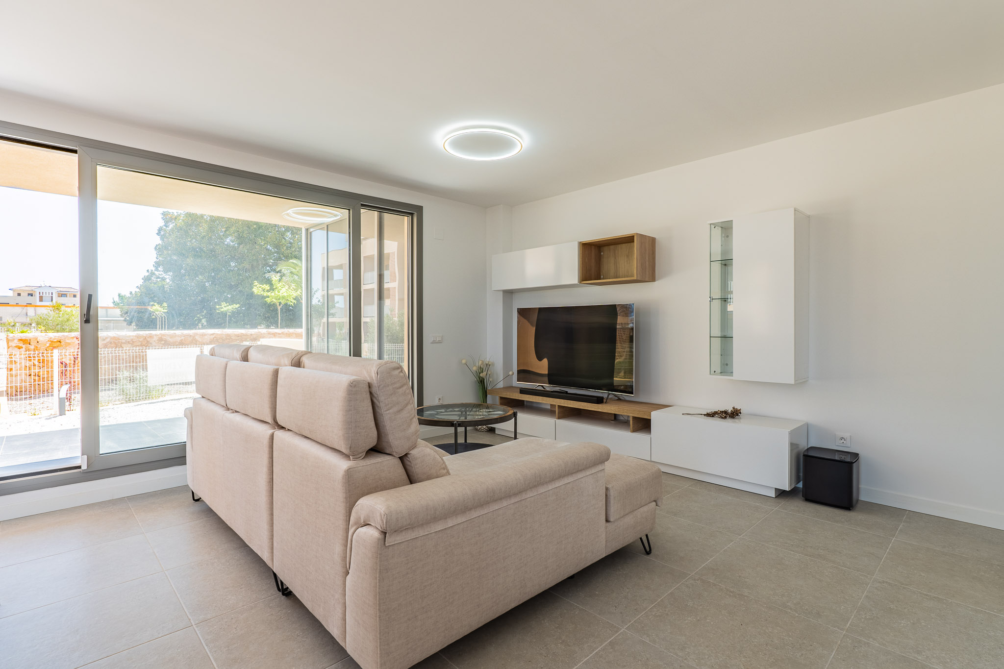 Bright living room featuring a beige sectional sofa, light grey tiled floor, and large sliding glass doors leading to the outdoor area. Modern media console with white and wood shelving.