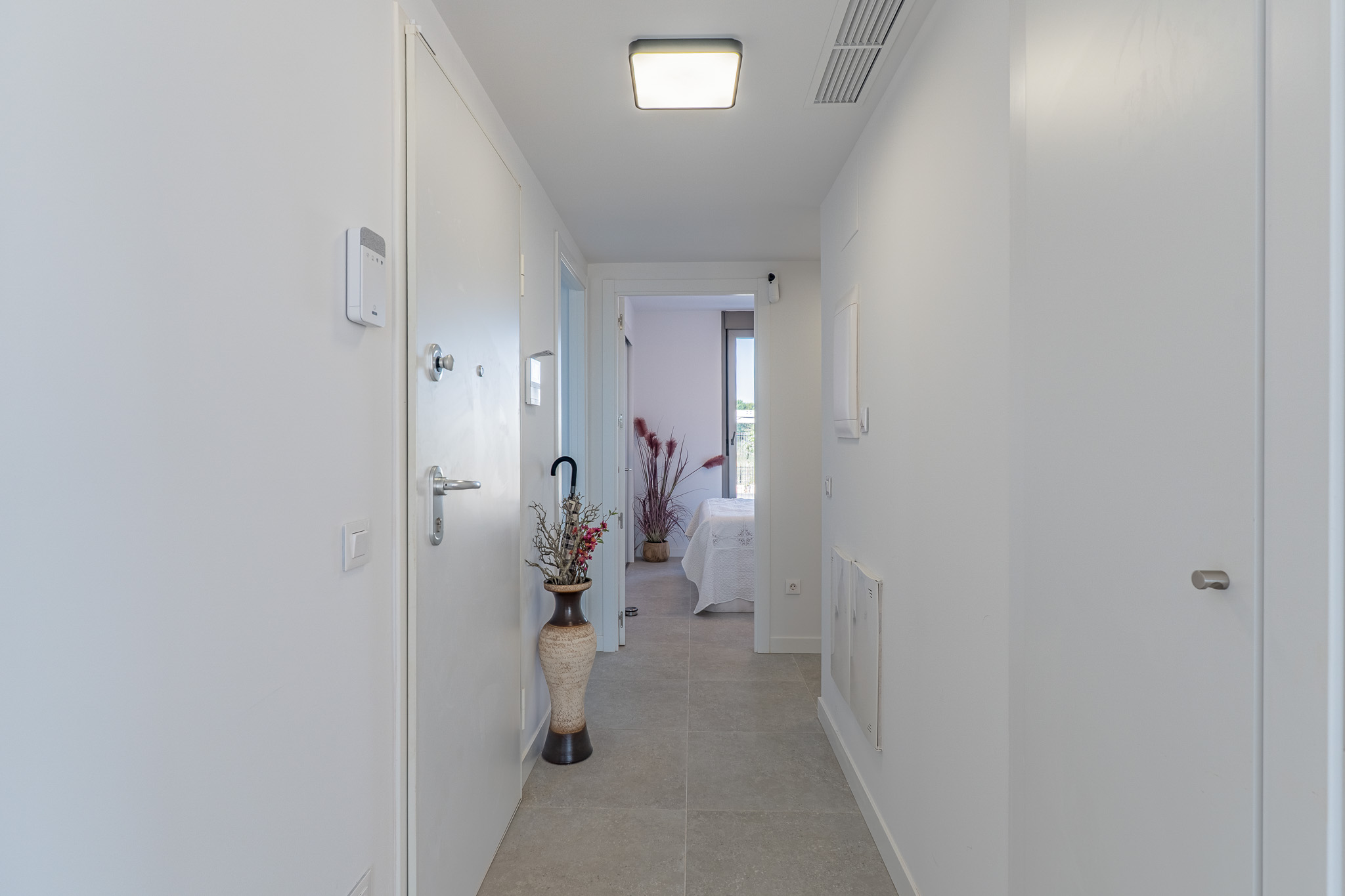 Bright, modern hallway with light grey tiled flooring and white walls. Features include a security panel, a contemporary ceiling light, and a glimpse into a bedroom.