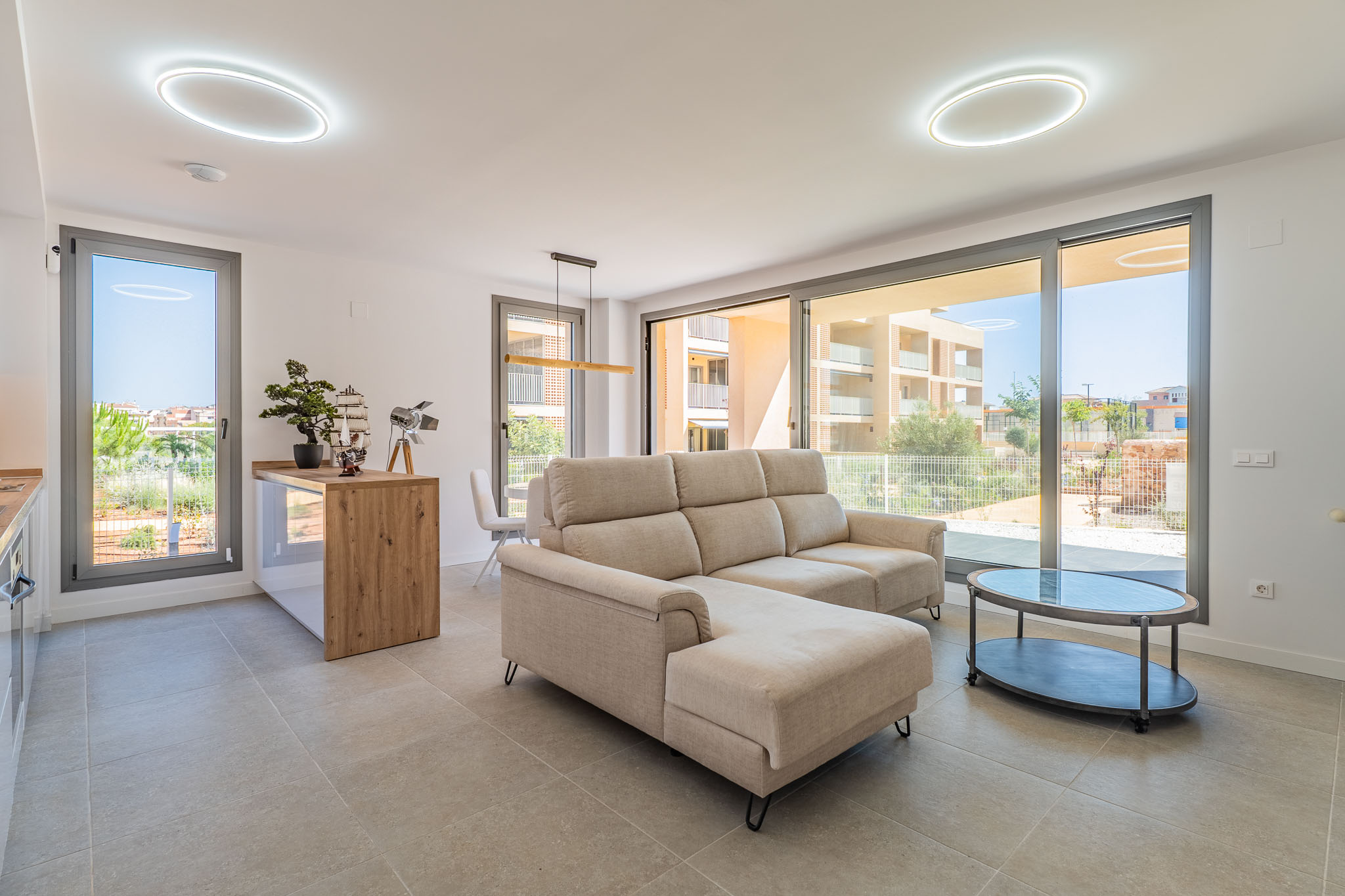 Bright open-plan living-dining area with a beige sectional sofa and oval coffee table. Large glass doors provide abundant natural light and residential views. Features an open kitchen with a wooden counter.