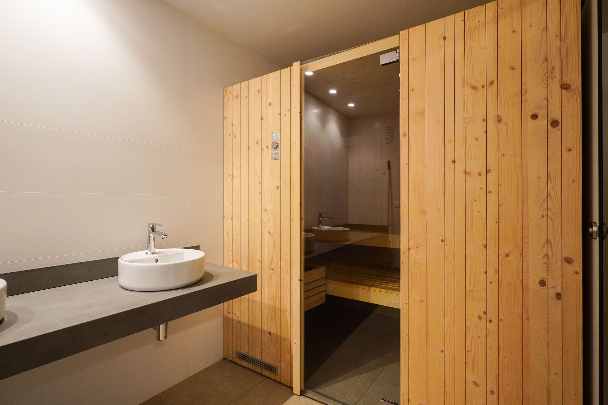 Changing area featuring a sleek floating vanity with a vessel sink and the inviting wooden-slatted sauna door with a glass panel, part of the communal social center.