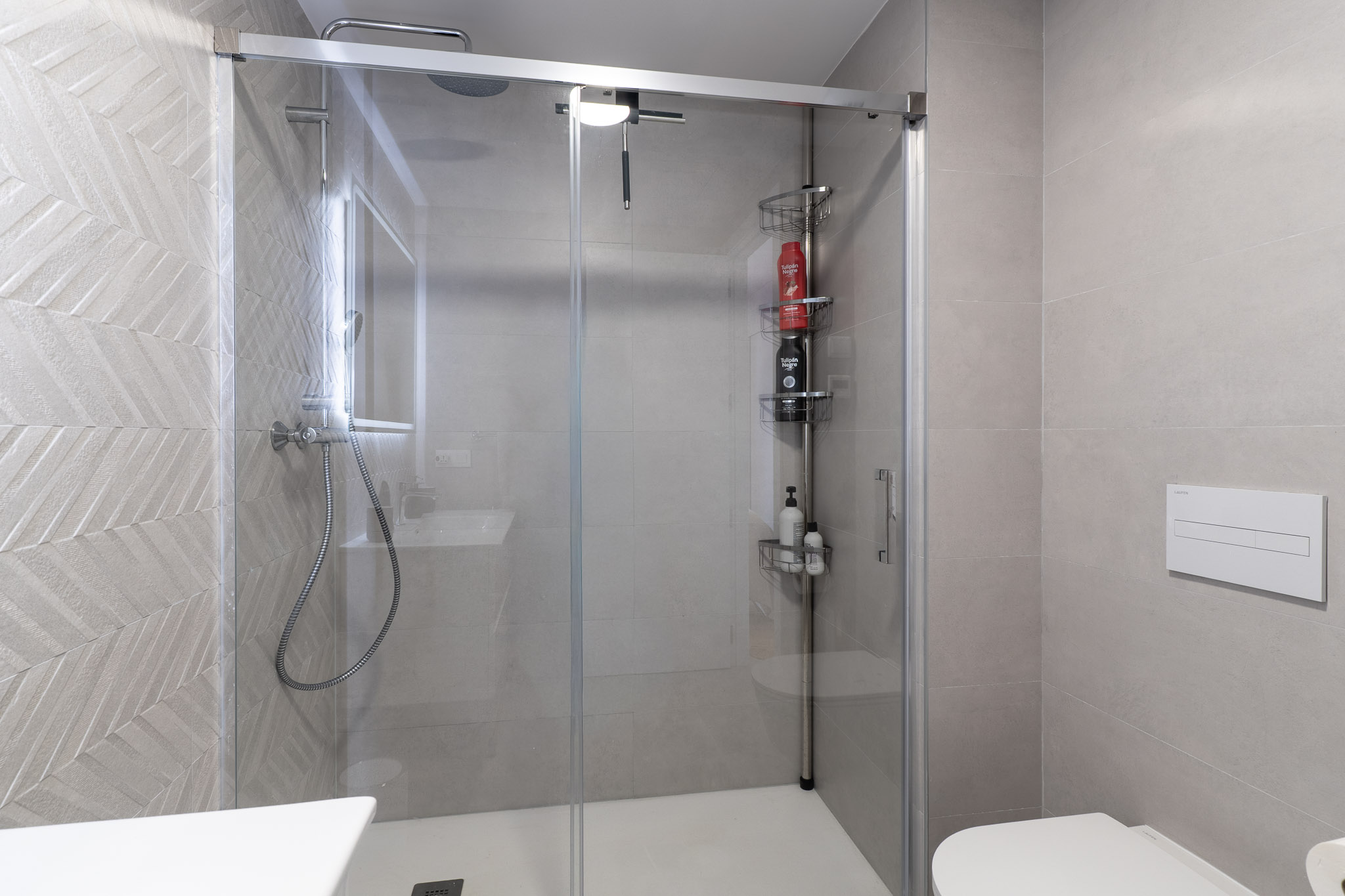 Contemporary bathroom featuring a glass shower enclosure and white tray. Light grey tiles with a distinct herringbone pattern adorn the shower wall, complemented by a rain shower and wall-mounted toilet.
