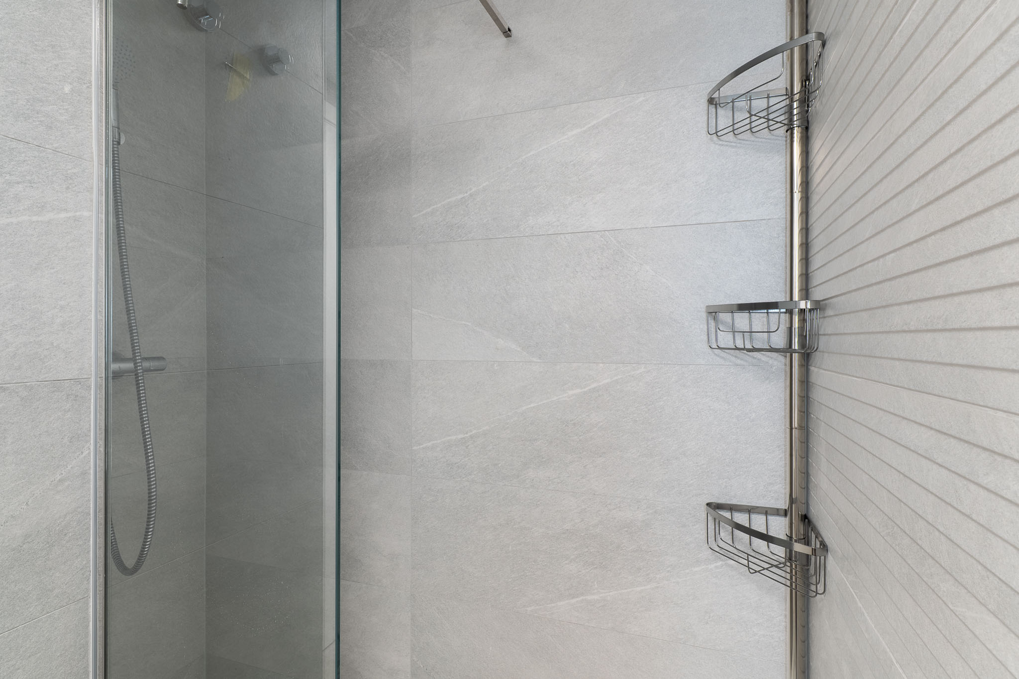 Contemporary shower area with large format light grey tiles, a textured accent wall, clear glass screen, and a practical chrome corner storage unit.