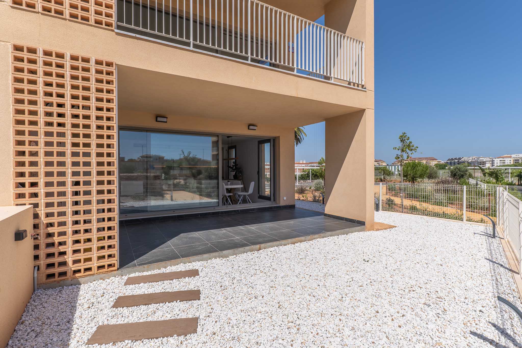 Covered ground-floor terrace with dark grey tiles, directly accessing a private white gravel garden. Large glass doors reveal a bright interior dining space, complemented by a modern decorative brick facade.