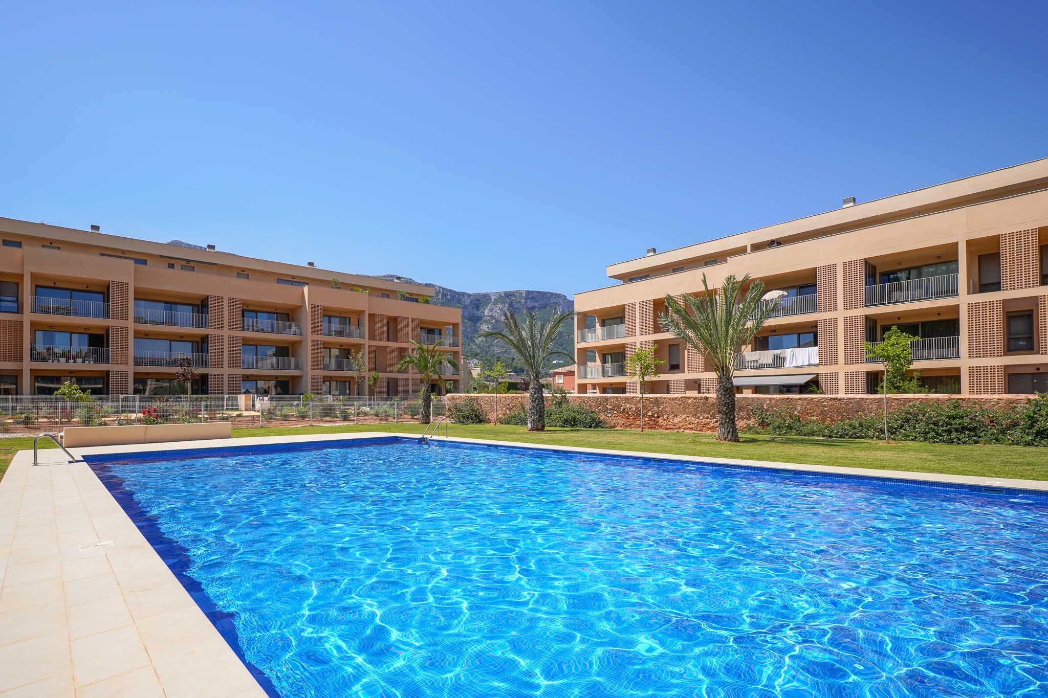 Expansive communal swimming pool with blue mosaic tiles, bordered by green lawn and modern residential buildings featuring balconies. Palm trees and mountains are visible in the background.