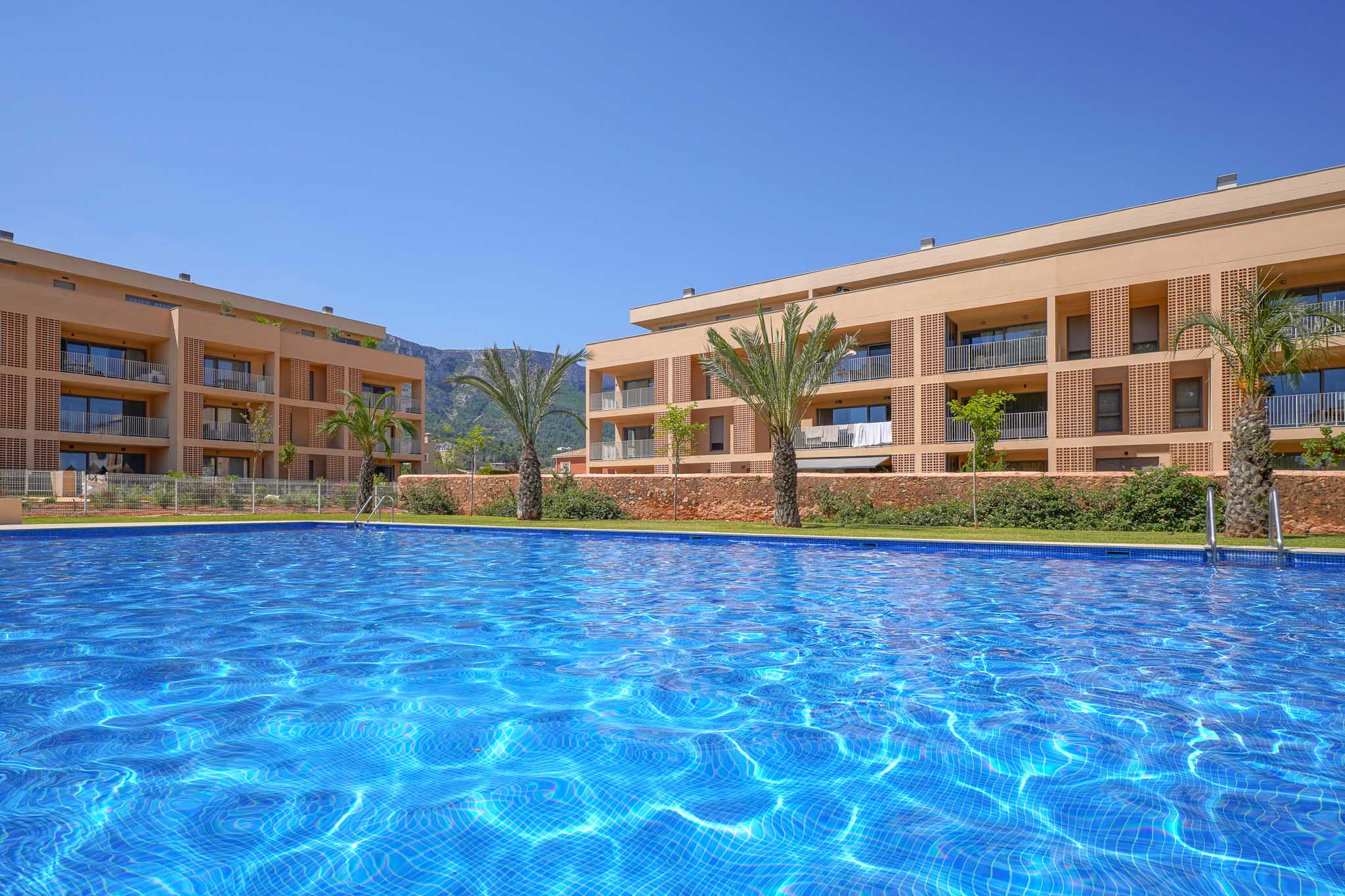 Expansive communal swimming pool with shimmering blue mosaic tiles, set against modern residential buildings featuring balconies and a distant mountain range under a clear sky.