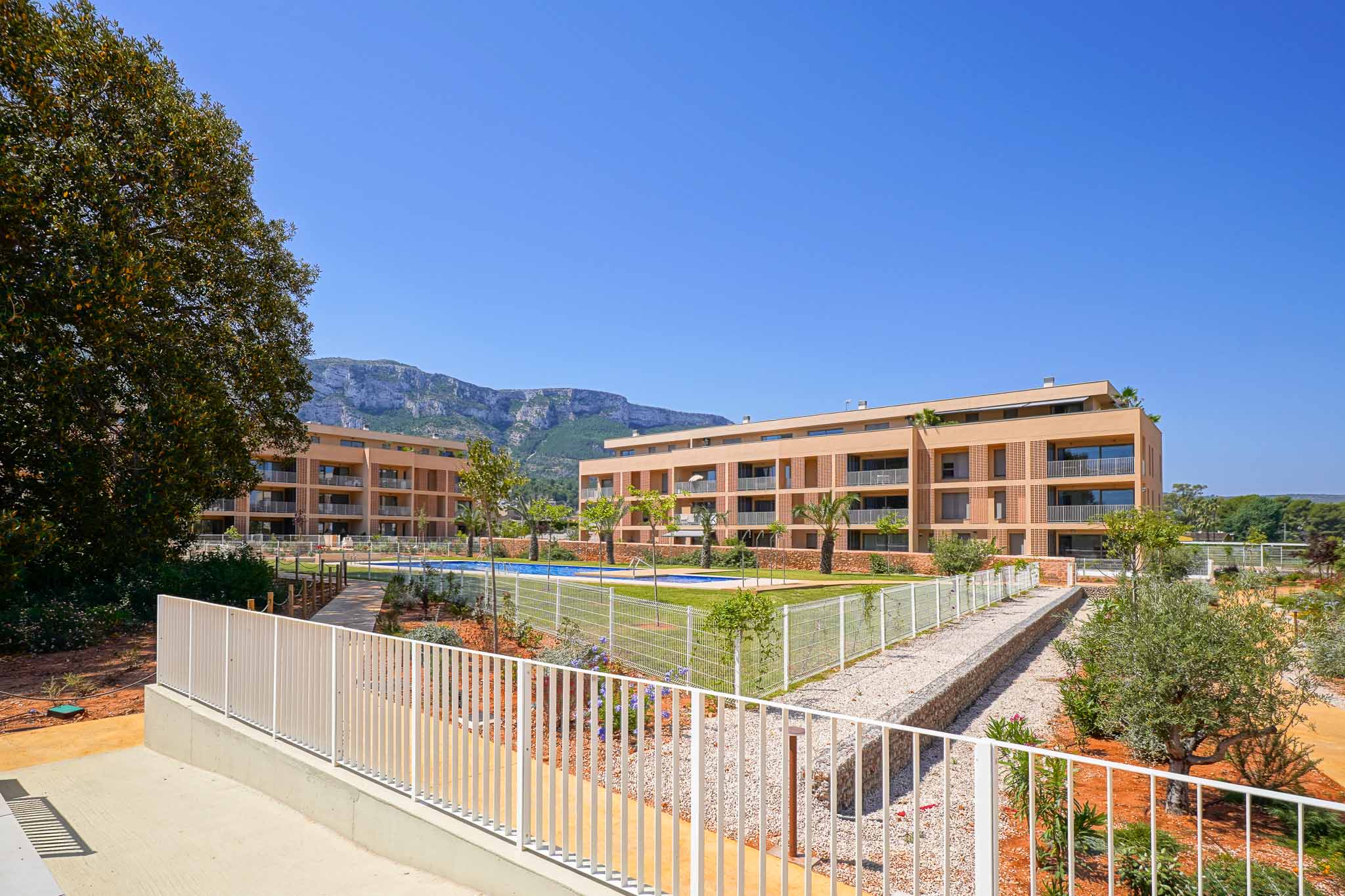 Exterior view of the modern residential complex featuring two light-colored buildings, a communal swimming pool, and landscaped gardens. Mountains are visible in the background.
