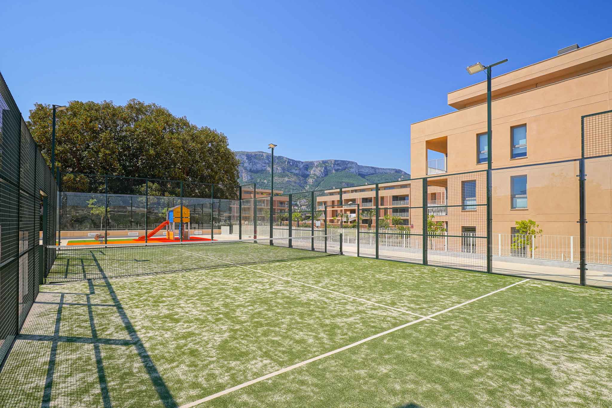 Green artificial turf padel court, fenced and equipped with lighting poles, adjacent to a children's playground and modern residential buildings with mountain views.