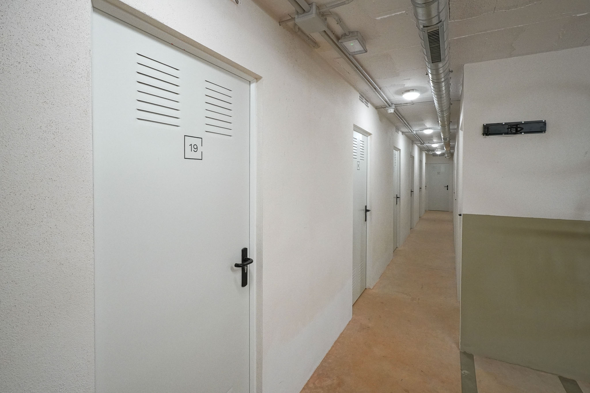Interior corridor featuring a series of white storage unit doors with ventilation slats and black handles. Exposed ceiling ducts and recessed lighting illuminate the concrete floor.