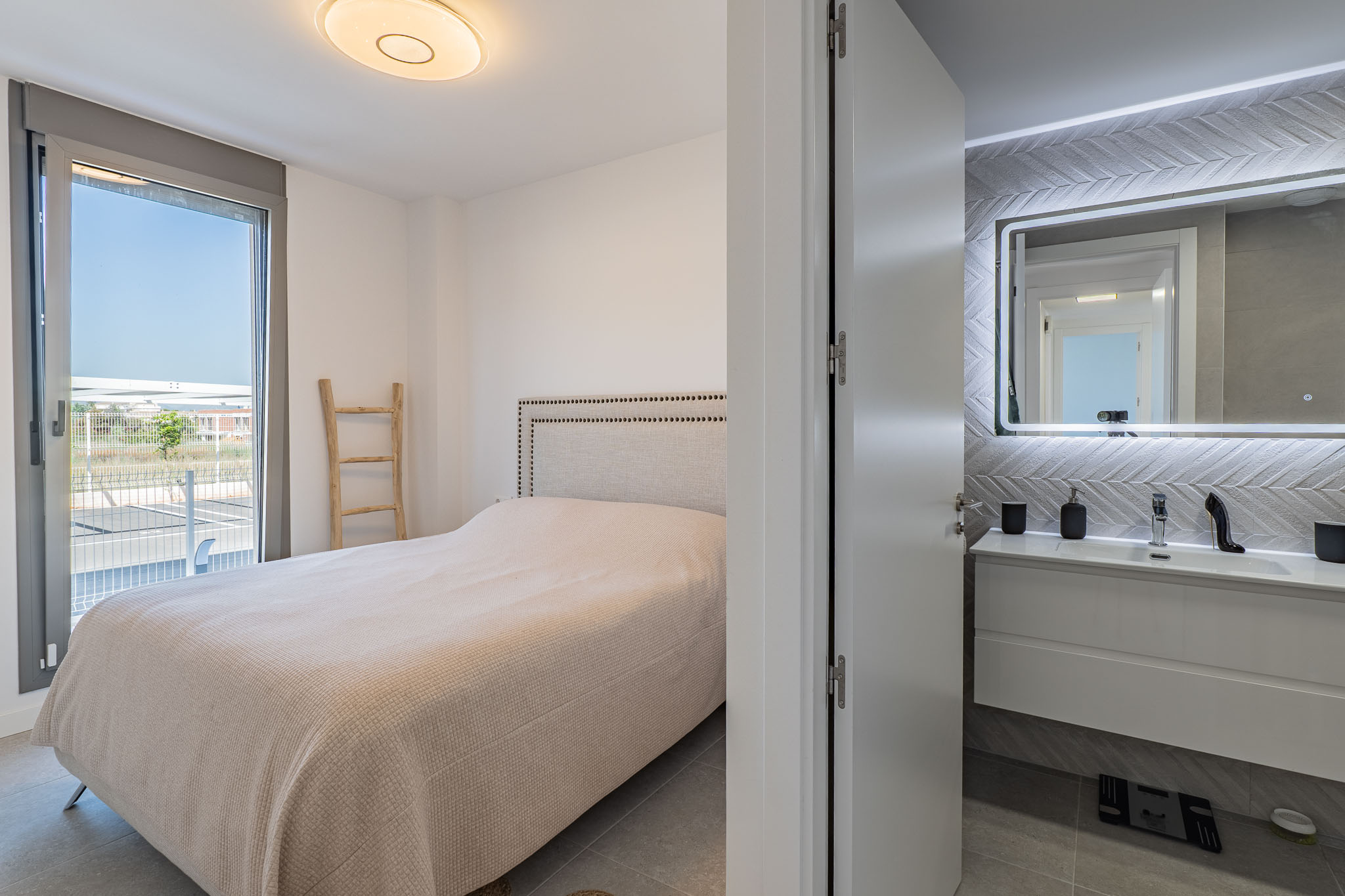 Main bedroom featuring a padded headboard bed and direct access to an outdoor terrace. An en-suite bathroom with a floating vanity and backlit mirror is visible.