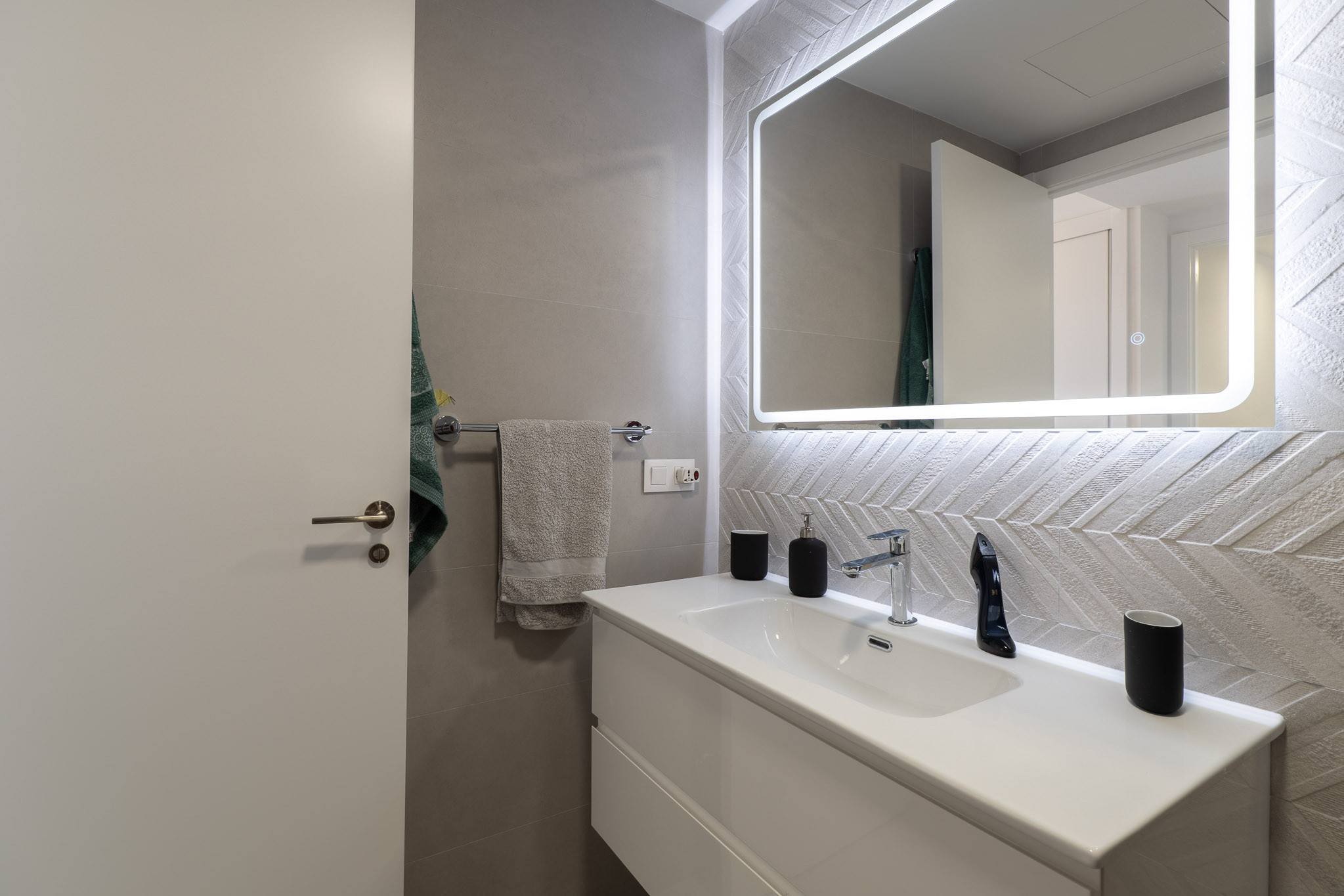Modern bathroom featuring a white floating vanity, chrome faucet, and an illuminated mirror. Accentuated by textured chevron wall tiles.